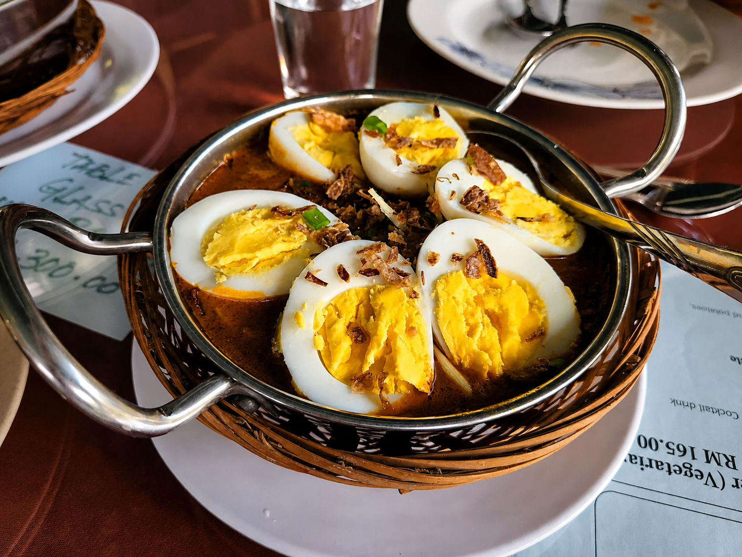 Halved boiled eggs in a dark spiced gravy, scattered with fried shallots, in a karahi set in a wicker holder.