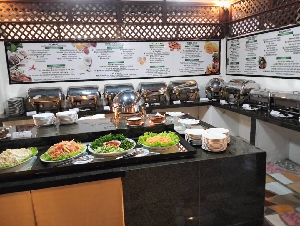 A long granite counter set with covered chafing dishes, stacked plates and wide platters of shredded salad, with framed boards mounted on the wall behind it.